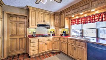 Une cuisine avec des meubles de rangement en bois, une cuisinière à gaz, un évier et une fenêtre ornée d’un rideau rouge.