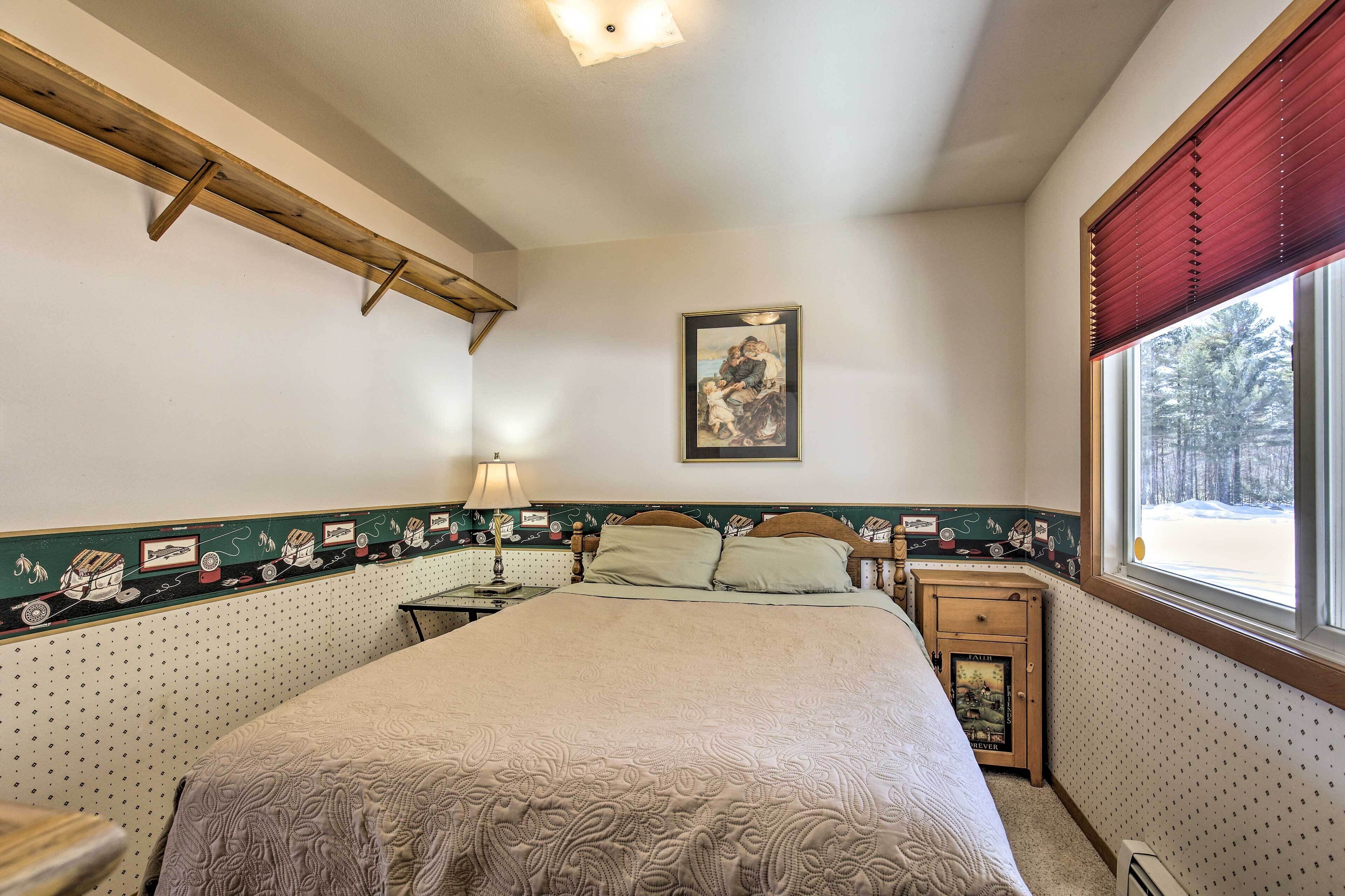 Une chambre à coucher avec un lit, une table de chevet, une lampe, un tableau au mur et une fenêtre donnant sur des arbres.