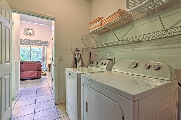 A laundry room with a front-loading washer and dryer, a drying rack above, and a door leading to a living area with a sofa and window.