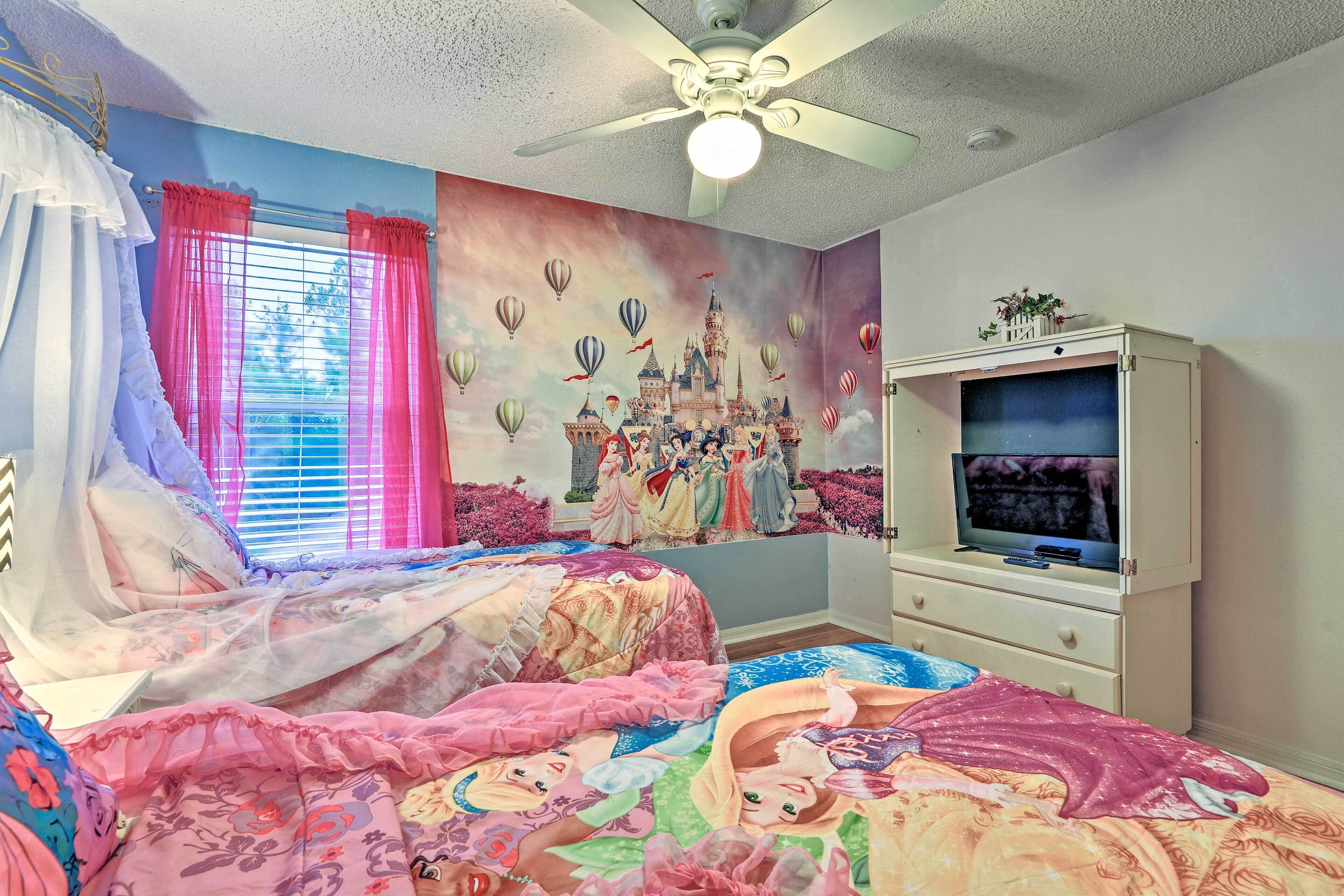 Ein Schlafzimmer mit einem Bett, das mit farbenfroher Bettwäsche bezogen ist, einem Deckenventilator, einem Fernseher auf einer Kommode und einem Wandgemälde mit einer Burg und Heißluftballons.
