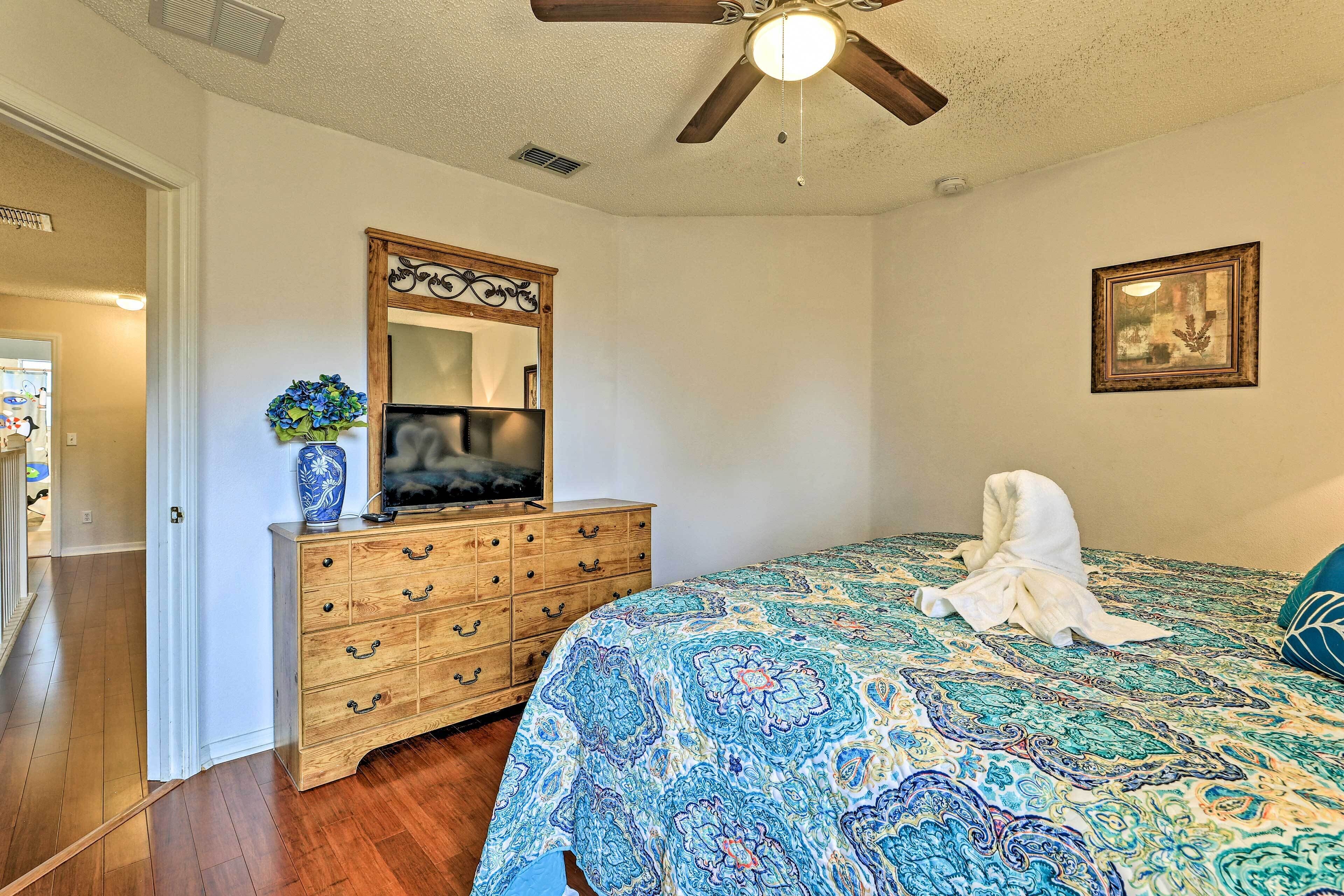 Une chambre avec une commode en bois, un grand lit recouvert d’une couvre-lit à motifs, un ventilateur de plafond et un tableau encadré accroché au mur.