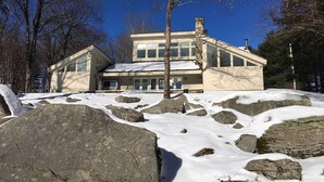 A large, modern house with a stone chimney, surrounded by snow-covered rocks and trees.