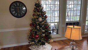 Ein Weihnachtsbaum mit Verzierungen und Lichtern, eine Wanduhr und eine Lampe in einem Raum mit Holzboden und einem Fenster.
