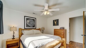 A bedroom with a bed, a ceiling fan, a nightstand, a lamp, a framed artwork, a wooden dresser, and a door. - Hot Tub & Patio: Pet-friendly Cabin in Munds Park! (Munds Park)