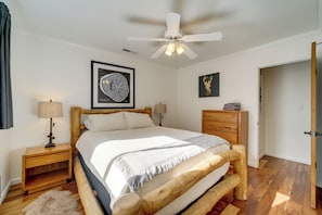 A bedroom with a bed, a ceiling fan, a nightstand, a lamp, a framed artwork, a wooden dresser, and a door.