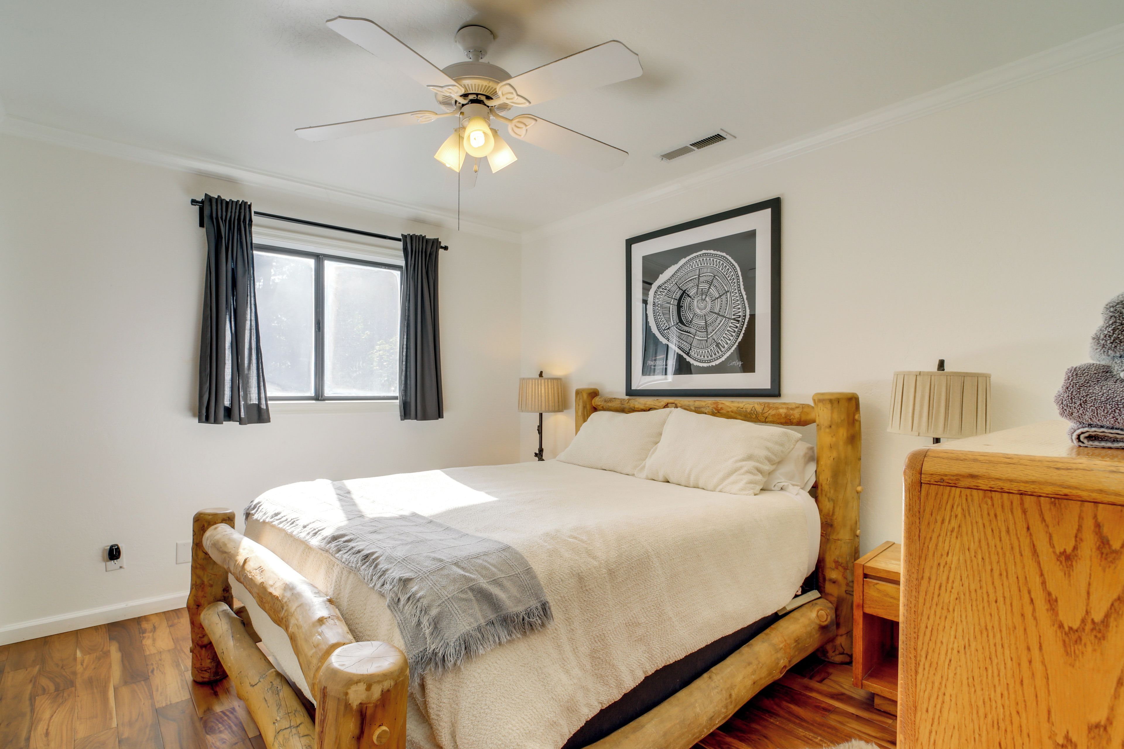 A bedroom with a wooden bed, a ceiling fan, a window with curtains, a bedside lamp, and a framed artwork on the wall.