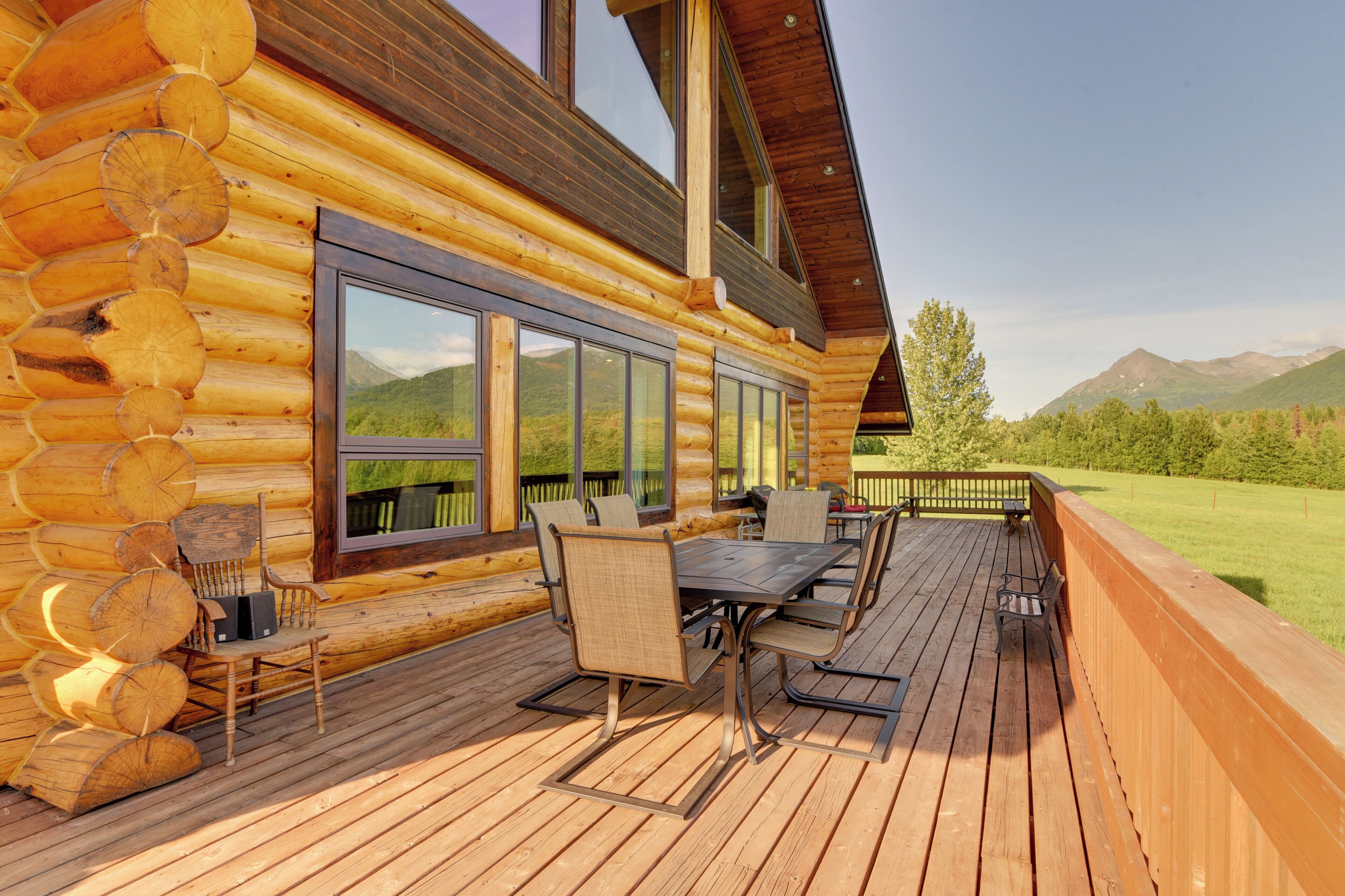 A log cabin with a deck featuring a table and chairs, large windows, and a view of mountains.
