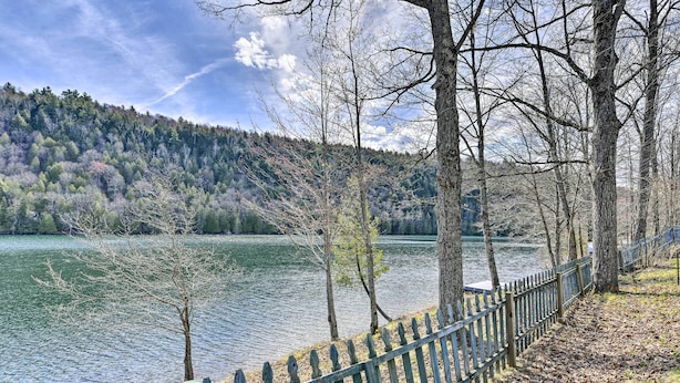 Blick auf einen See mit einer Holzlattenveranda, Bäumen und einem bewaldeten Hügel im Hintergrund.