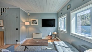 Ein gemütliches Wohnzimmer mit Holzbalkendecke, einem an der Wand befestigten Flachbildfernseher, einem großen Fenster mit Seeblick und einer komfortablen Sitzecke mit Sofa und Couchtisch.