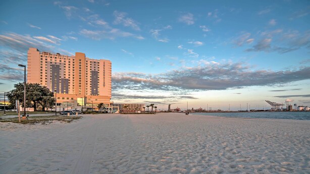 A beachfront hotel with a sandy beach, a clear sky, and a distant view of industrial structures. - Charming Gulfport Getaway Only 3 Blocks to Beach! (Gulfport)