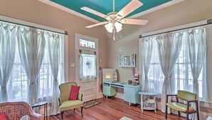A room with a teal ceiling, white curtains, a ceiling fan, a green armchair, a small table, and a desk with a mirror and a shelf. - Charming Gulfport Getaway Only 3 Blocks to Beach! (Gulfport)