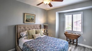 Une chambre à coucher avec une tête de lit capitonnée, une couvre-lit à motifs, un ventilateur de plafond, une table de chevet avec une lampe et une fenêtre avec des stores.