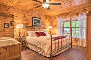 A wooden cabin bedroom with a bed, two bedside lamps, a ceiling fan, a framed picture on the wall, and a window with curtains. - Cabin w/ Deck + Fireplace < 3 Mi to Dollywood! (Pigeon Forge)