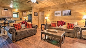 A cozy cabin interior with two leather sofas, a coffee table, wooden floors, a ceiling fan, and a kitchen area visible through an open door. - Cabin w/ Deck + Fireplace < 3 Mi to Dollywood! (Pigeon Forge)