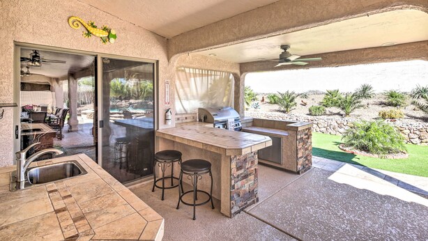 A covered outdoor kitchen area with a bar stool, a grill, and a view of a landscaped garden. - Heated Pool & Game Room: Lake Havasu Retreat (Lake Havasu City)