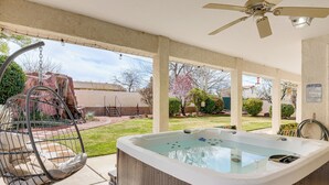A hot tub in a covered outdoor area with a ceiling fan, a hanging chair, and a grassy yard with trees and a building in the background.