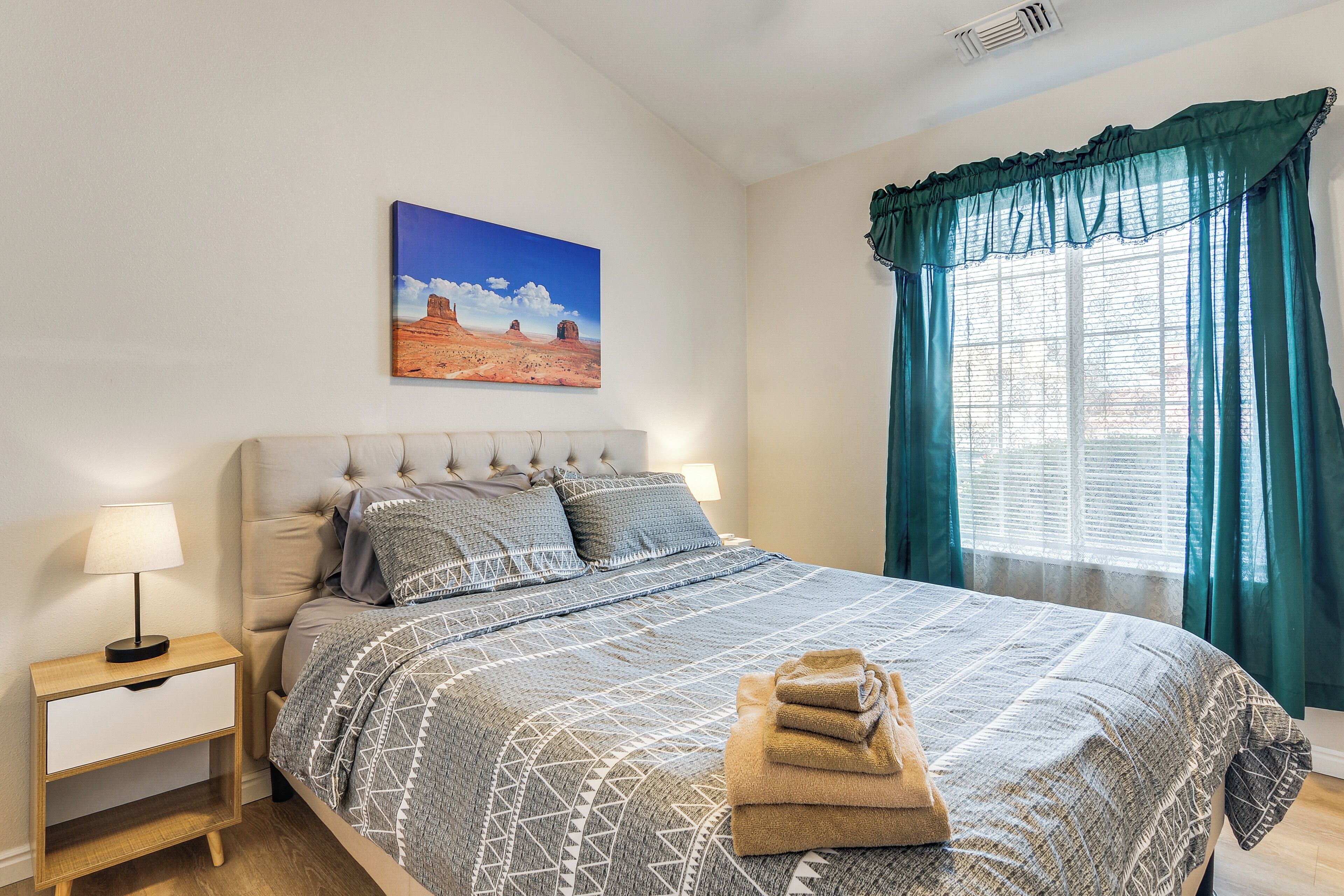 A bedroom with a bed, a nightstand, a lamp, a painting, and a window with curtains.