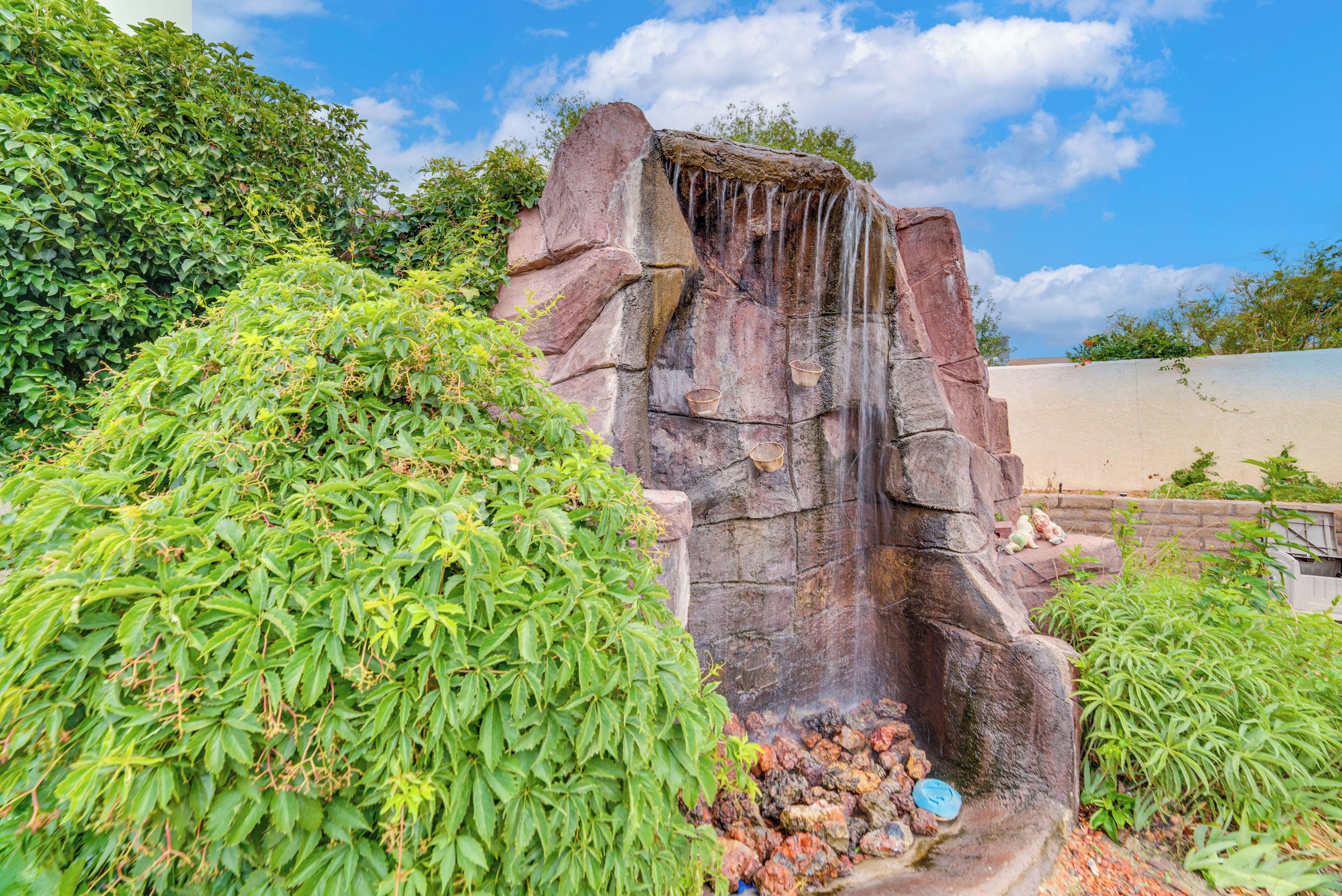 A rock waterfall with cascading water, surrounded by lush greenery and a clear blue sky.
