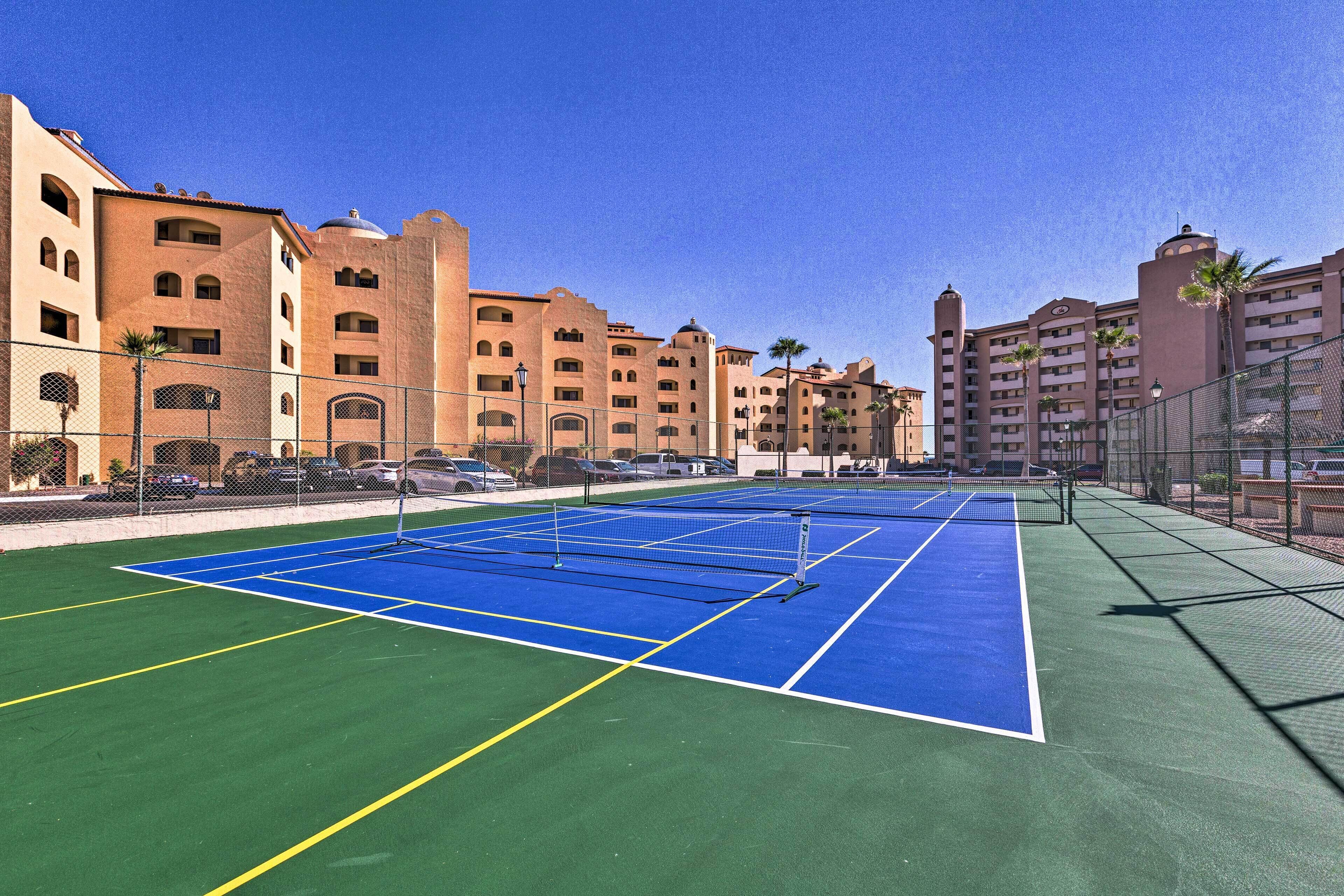 A tennis court with a net, surrounded by a fence, and adjacent to a building with multiple balconies and palm trees.