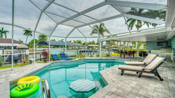 An indoor swimming pool with lounge chairs, a sunshade structure, and a view of palm trees. - Canalfront Cape Coral Home With Dock & Bbq! (Cape Coral)