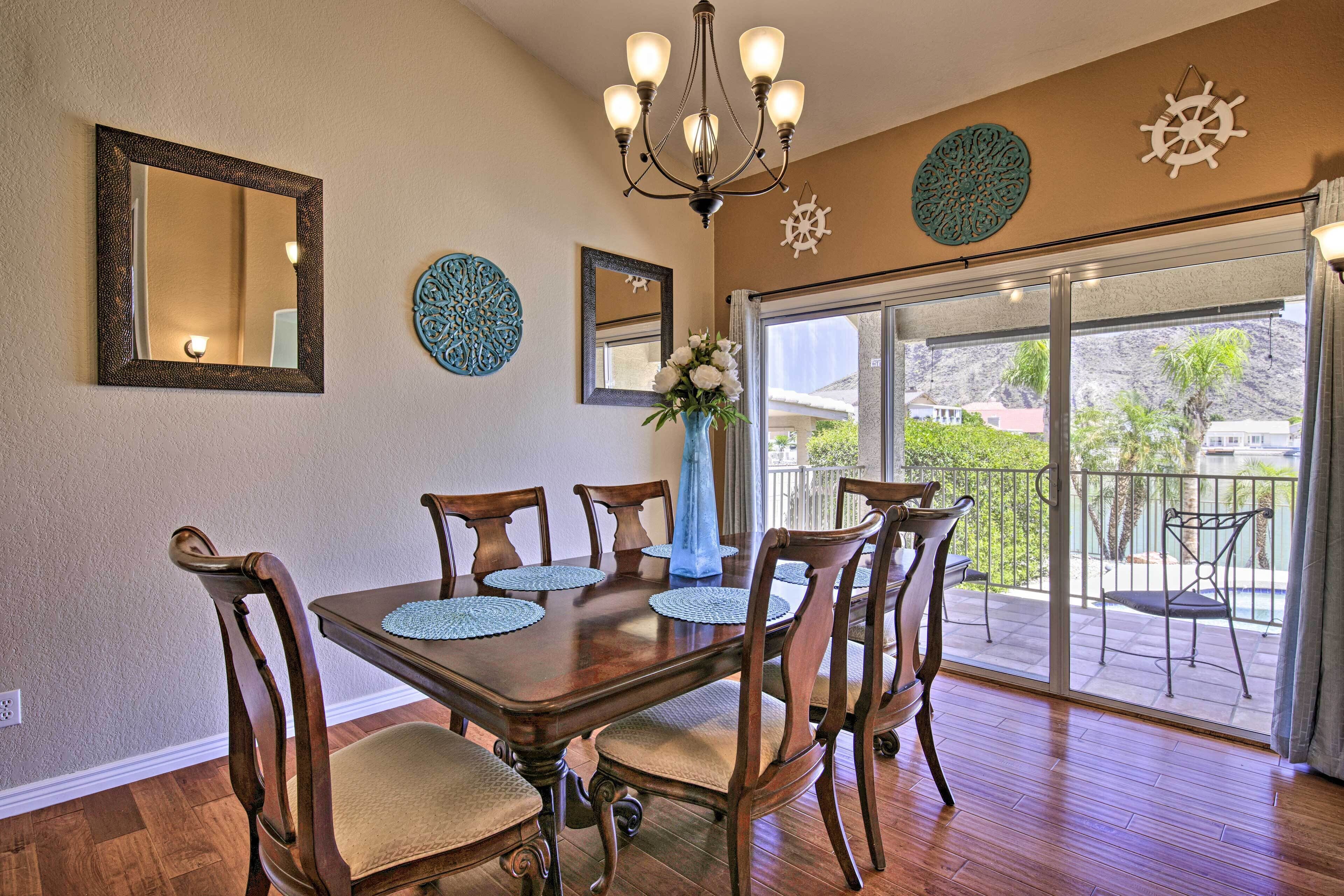 A dining room with a wooden table, chairs, and a chandelier. There are decorative wall mirrors and a large window with a view of the outdoors.