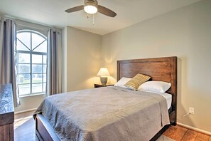 Quarto com cama, mesa de cabeceira, luminária, ventoinha no teto e janela com cortinas.