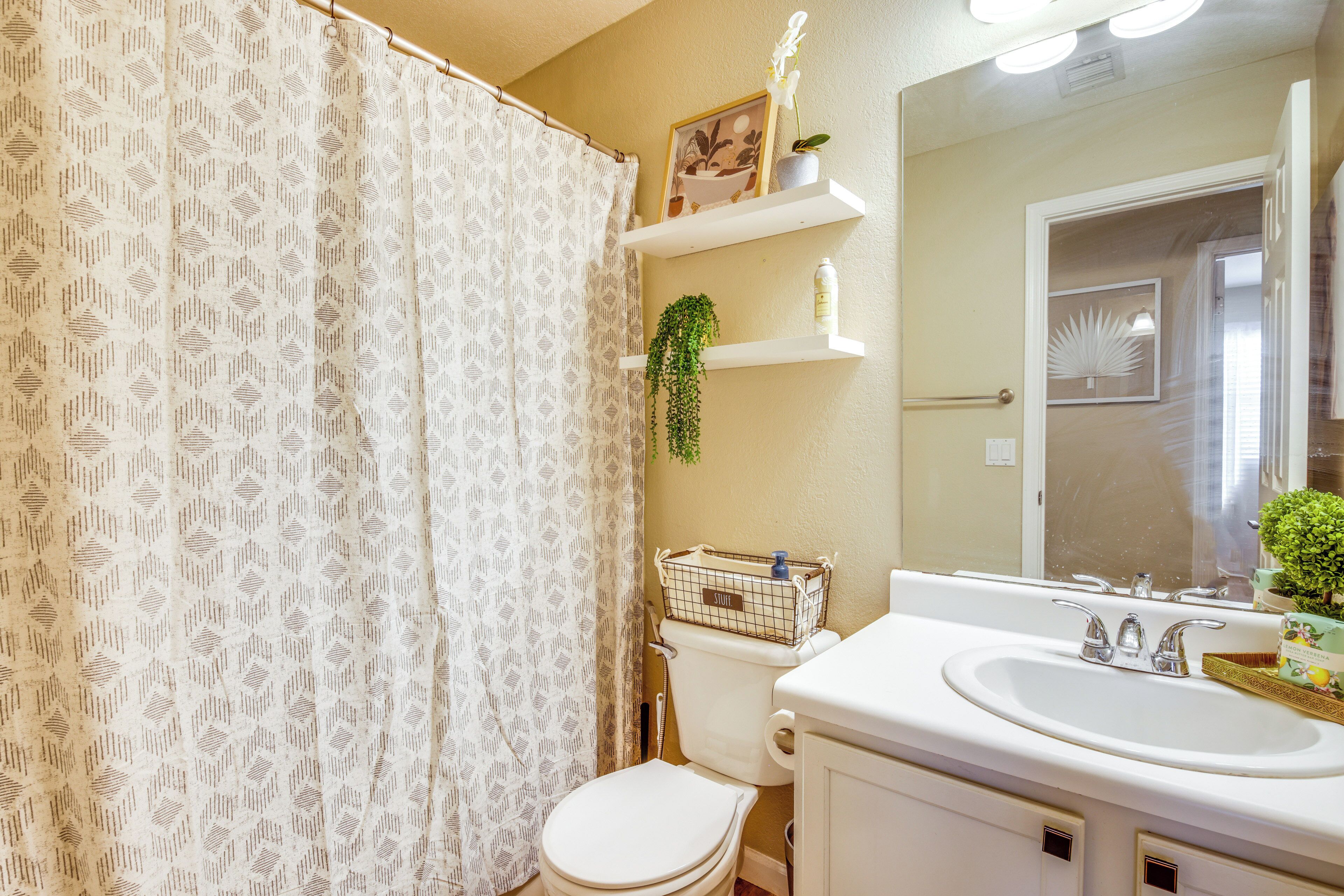 Baño con inodoro, lavabo y cortina de ducha. Hay repisas sobre el lavabo con artículos de aseo y una planta pequeña.