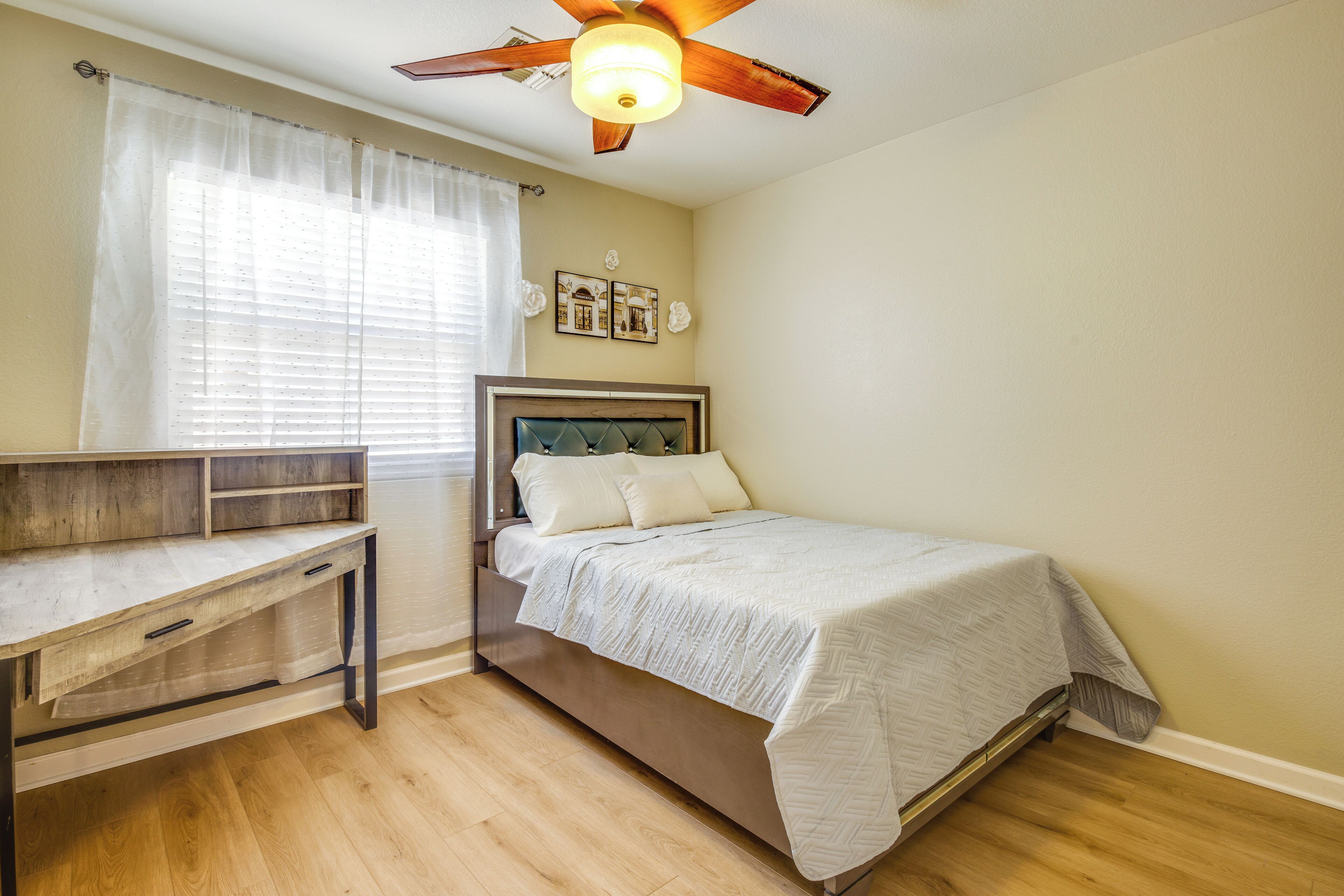 A bedroom with a bed, a desk, a ceiling fan, and a window with sheer curtains.