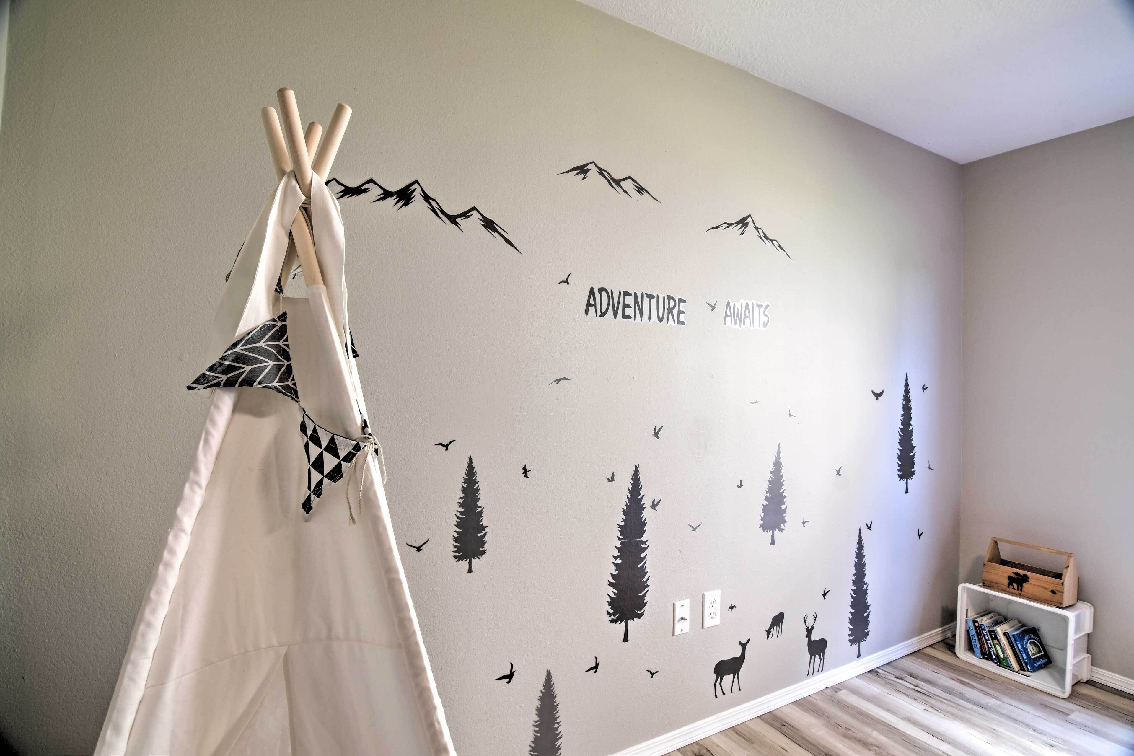 Ett rum med en vägg dekorerad med en väggdekor med naturmotiv, ett vitt tipi-tält och en liten bokhylla.
