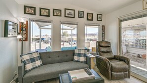 A living room with a gray sofa, a brown leather recliner, and a coffee table. There are framed pictures on the wall and a large window with a view of a parking lot. - Steps to Beach: Lake Havasu City Home w/ Grill! (Lake Havasu City)