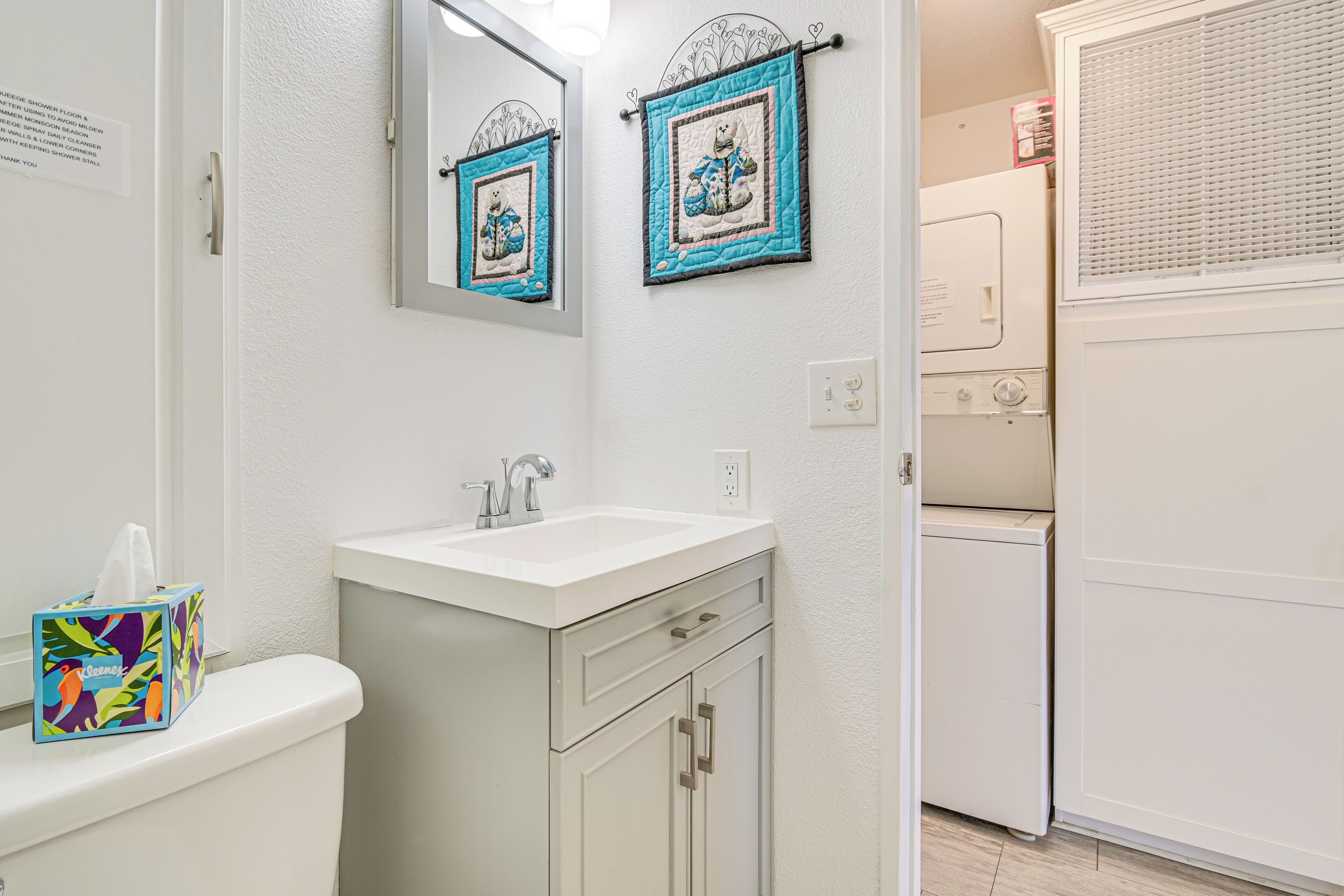 A bathroom with a white sink, mirror, and toilet. There is a towel rack with a teddy bear decoration and a box of tissues on the sink.