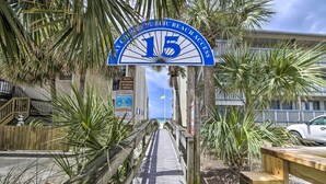 Ein hölzerner Steg führt zu einem Zugang zur Strandbucht, mit einem Schild, auf dem '15 Öffentlicher Strandzugang' steht.