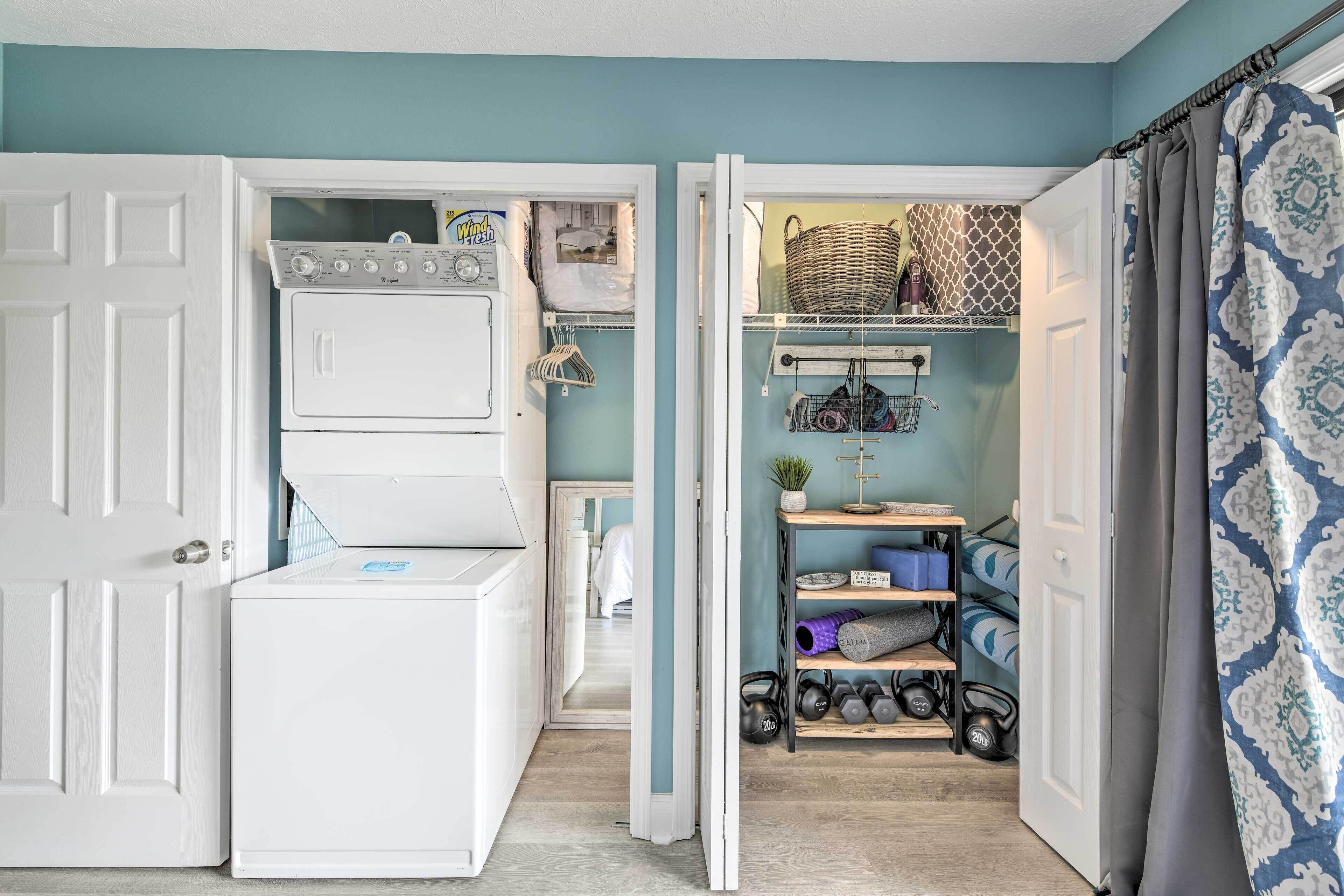 A laundry room with a front-loading washer and dryer, a shelf with baskets and towels, and a rack with exercise equipment.