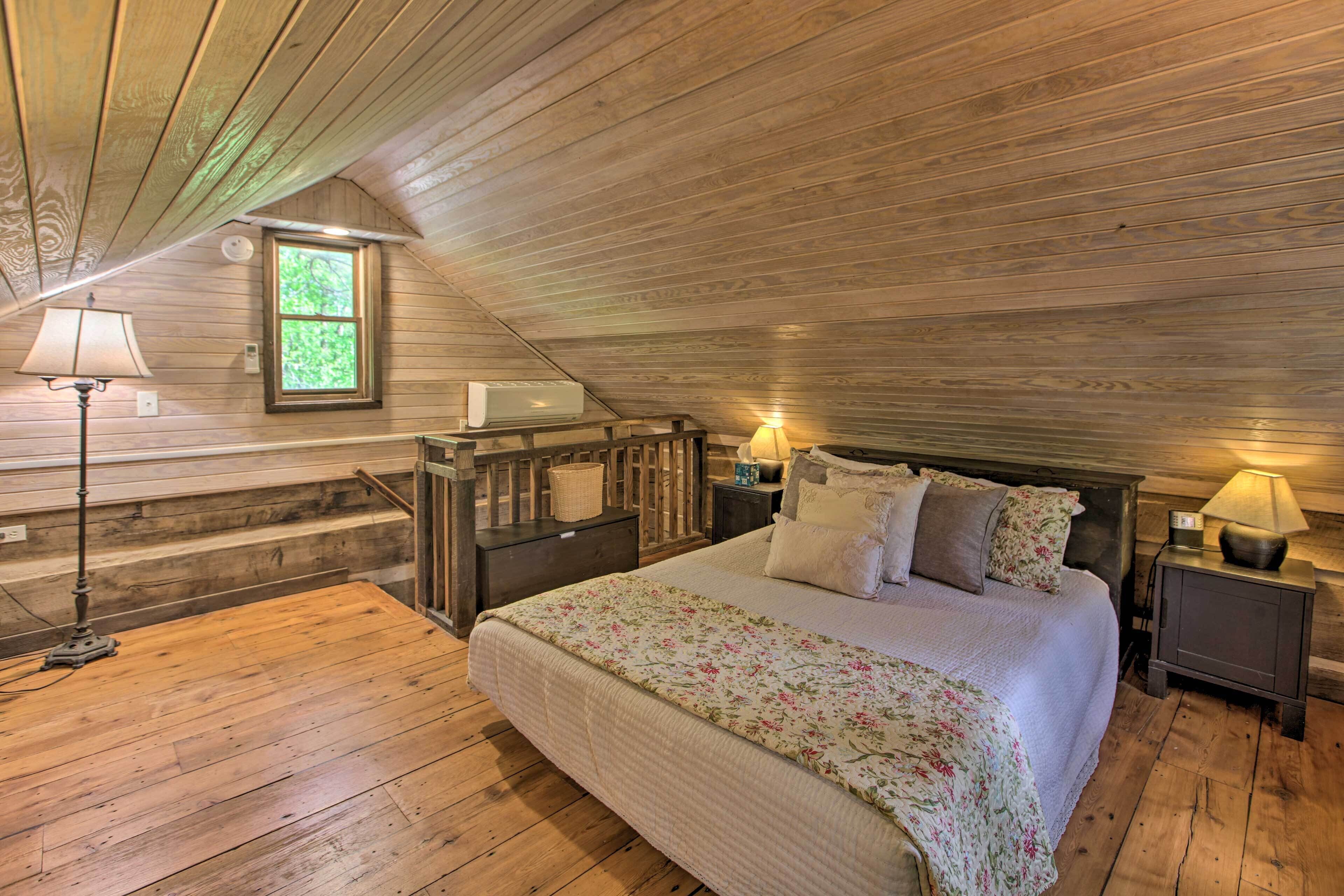 A cozy attic bedroom with a wooden ceiling, a large bed with floral bedding, two bedside lamps, and a window with a view of greenery.