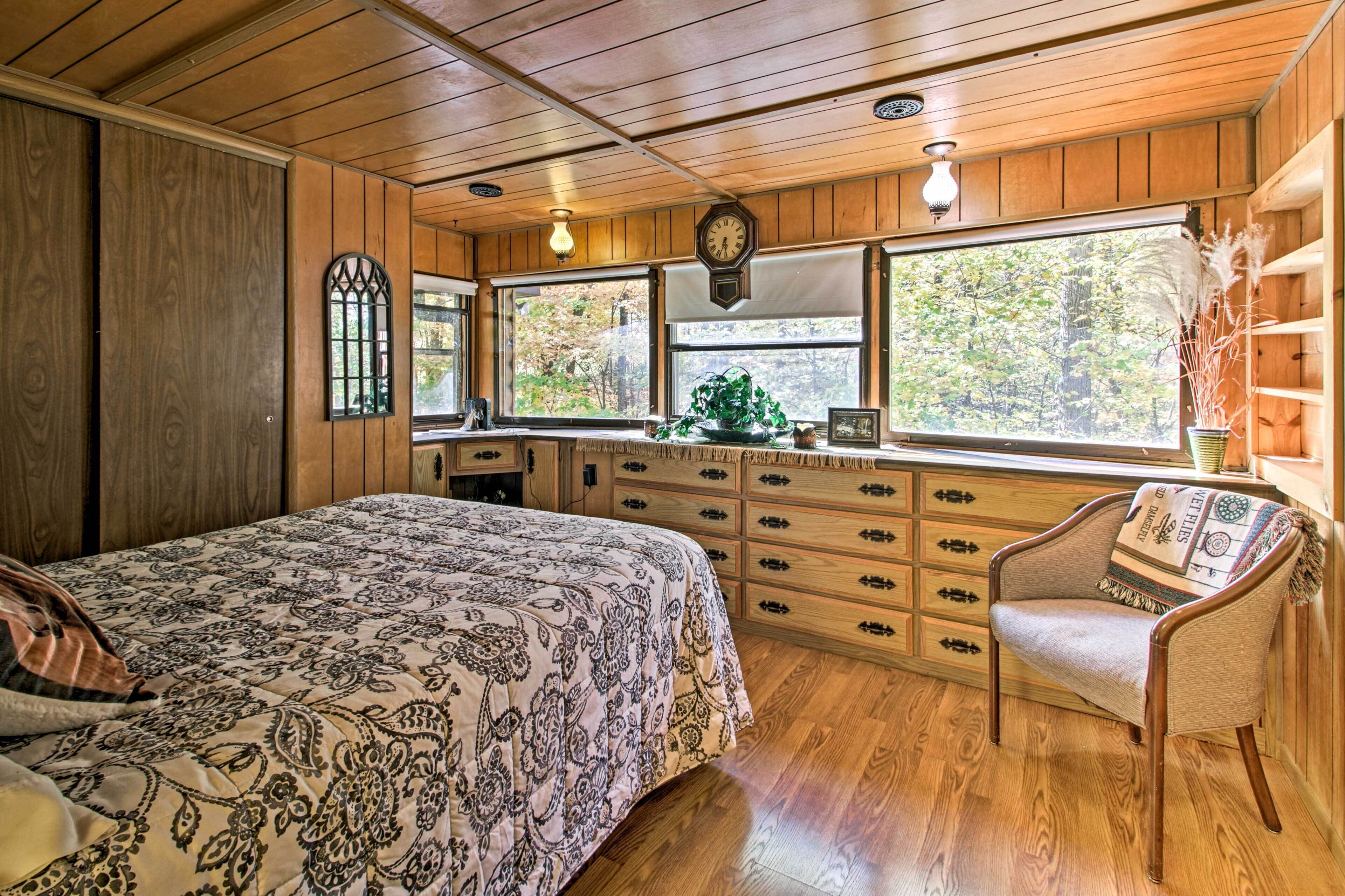 A wooden cabin bedroom with a bed, dresser, chair, and large windows.
