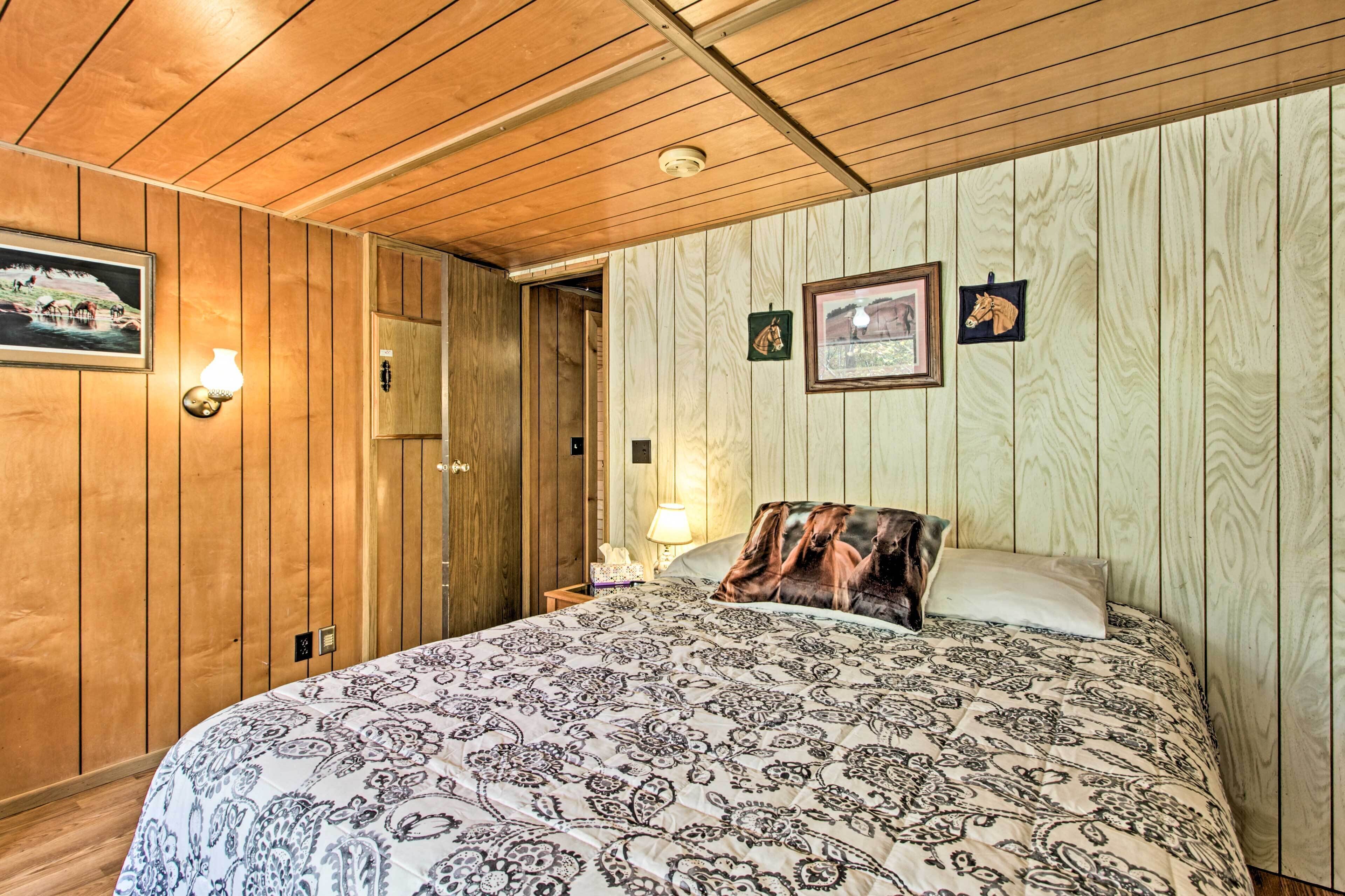 A bedroom with wooden panel walls, a bed with a patterned bedspread, a nightstand with a lamp, and framed pictures on the wall.