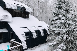 A snow-covered wooden cabin with a steep roof and multiple windows, surrounded by a snowy forest.