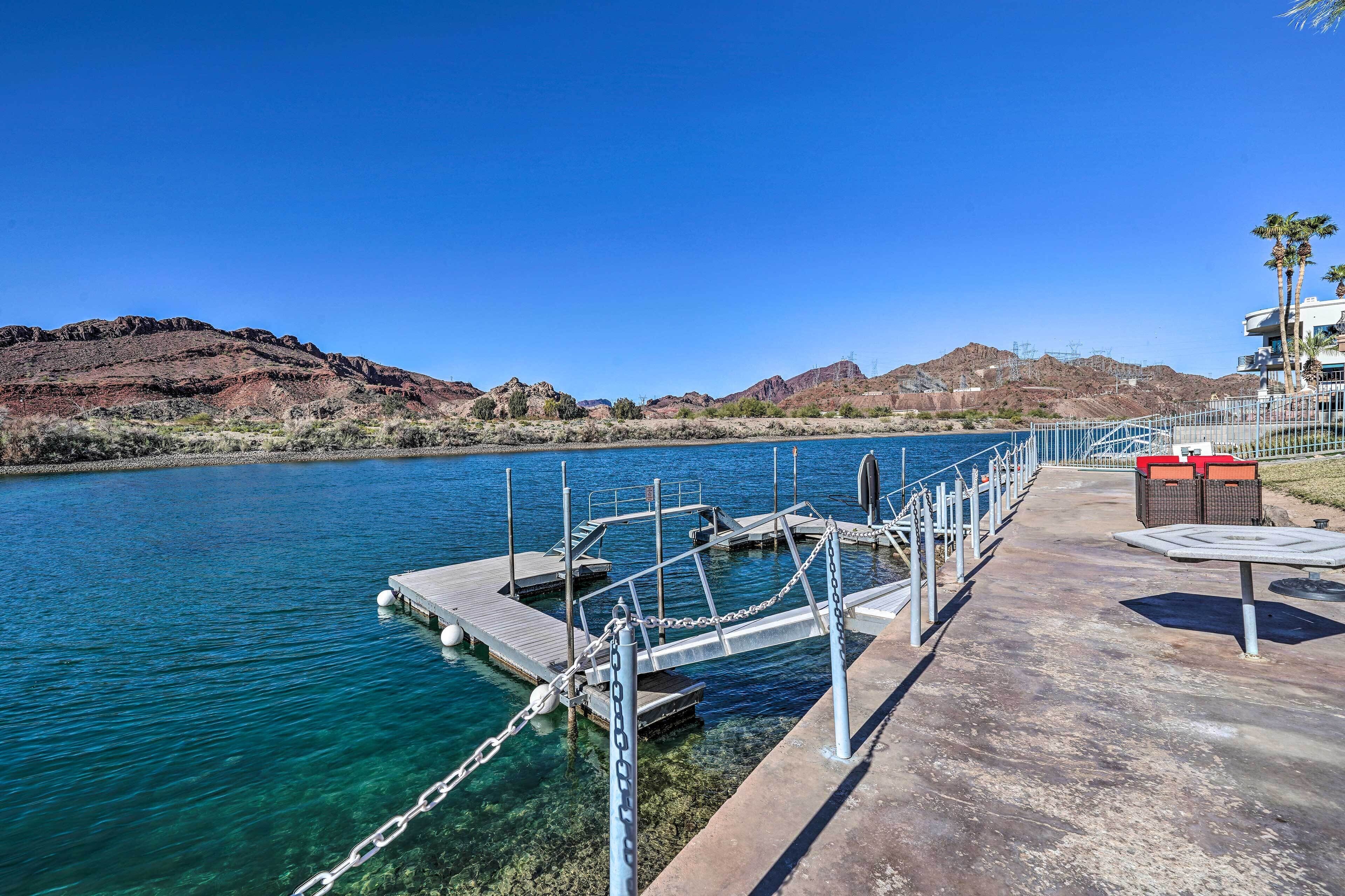 A clear blue waterfront with a dock, a concrete walkway, and a building with palm trees.