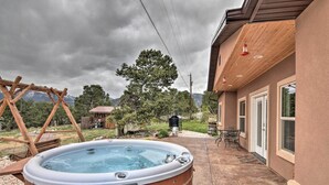 A hot tub in a backyard with a wooden deck, a patio with outdoor furniture, and a house with large windows and a covered porch. - Stunning Home w/ Fire Pit, 11 Mi to Mt Yale! (Buena Vista)