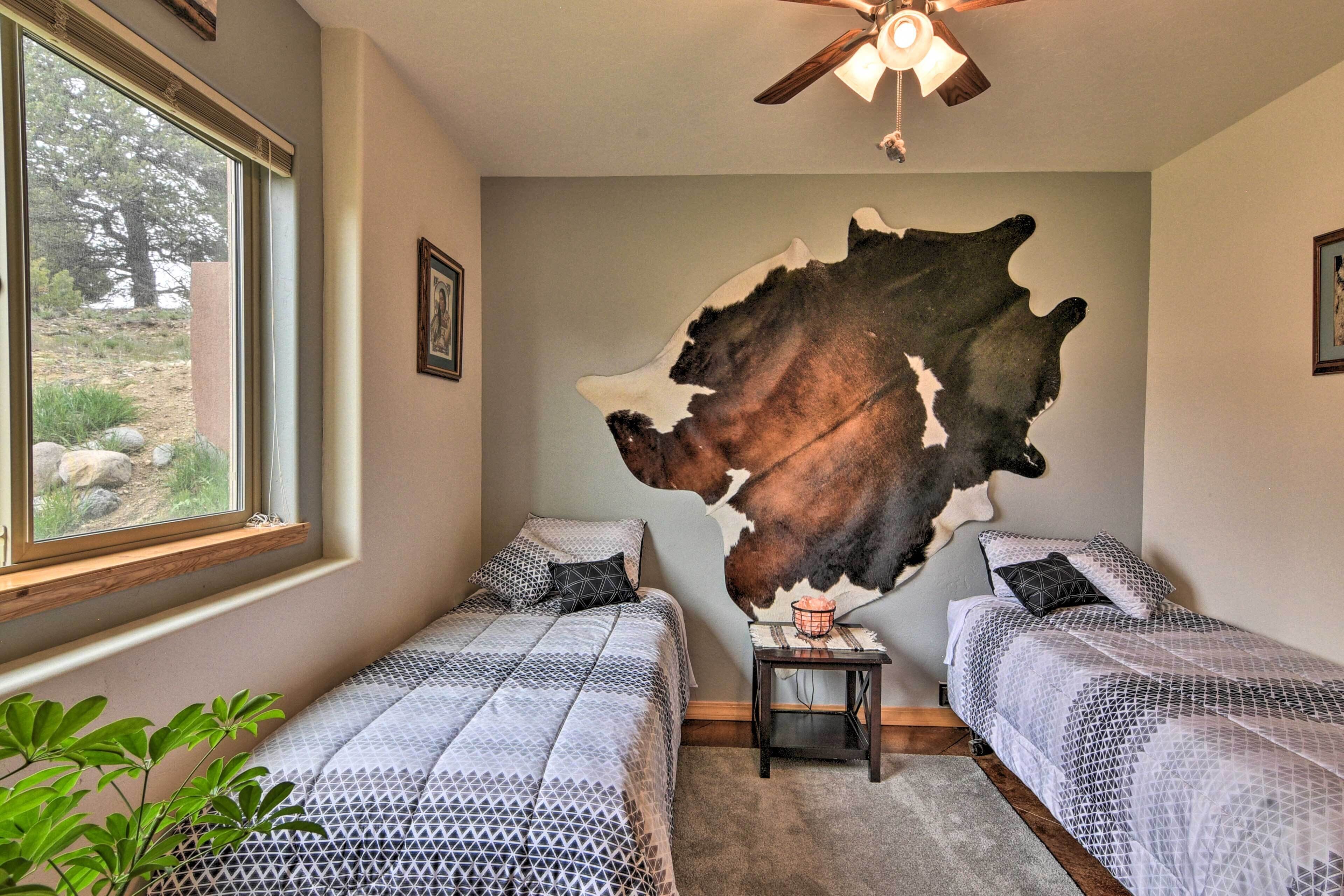 A bedroom with two beds, a large wall mural, a ceiling fan, and a window with a view of trees.