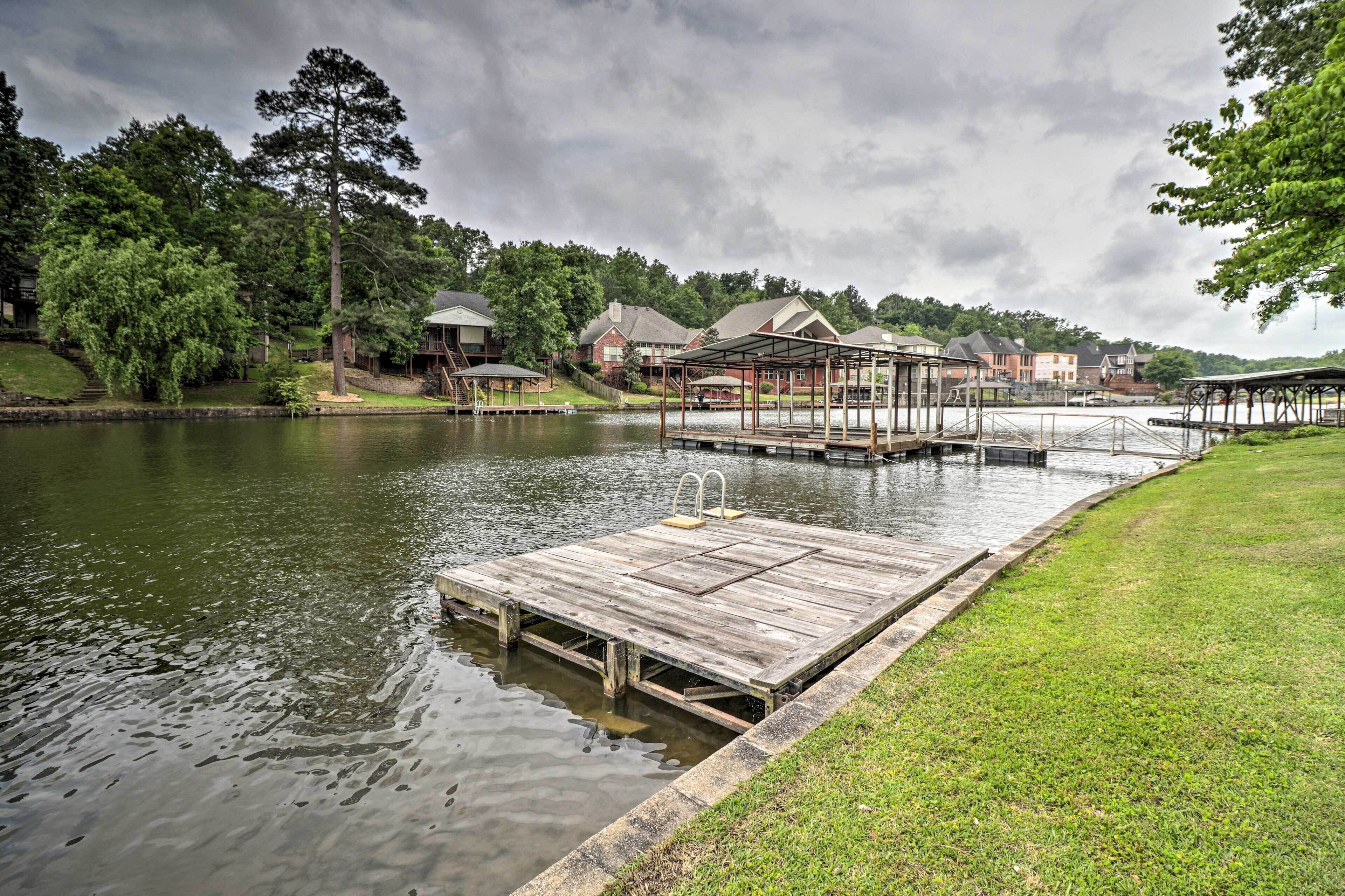Une jetée en bois s’étendant dans un lac paisible, entourée d’une végétation luxuriante et avec des bâtiments résidentiels en arrière-plan.