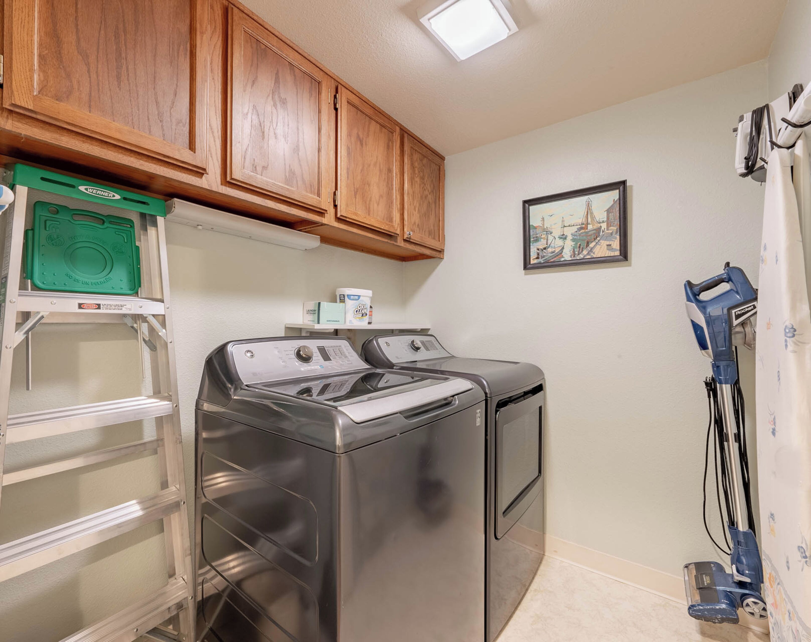A laundry room with two stainless steel washers, wooden cabinets, a green storage box, a framed picture on the wall, and a vacuum cleaner.