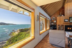 A room with large windows overlooking the ocean, wooden walls, and a fireplace.
