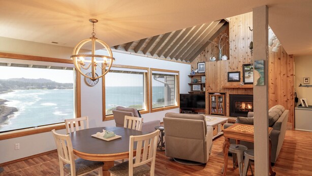 A cozy living room with a fireplace, wooden walls, a dining area with a view of the ocean, and a chandelier. - Idyllic Coastal Haven - Whale Watch in Depoe Bay! (Depoe Bay)
