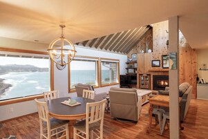 A cozy living room with a fireplace, wooden walls, a dining area with a view of the ocean, and a chandelier.