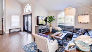 A spacious living room with a black leather sofa, a coffee table, and a TV. There is a large window with a view of greenery outside.