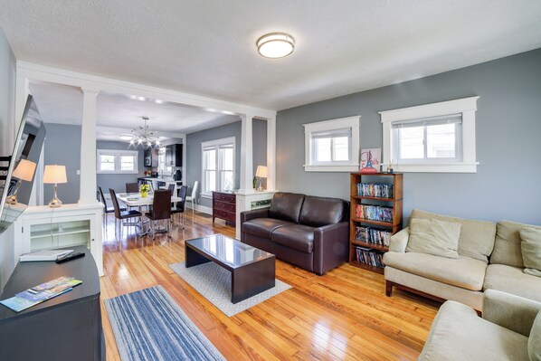 A spacious living room with a brown leather sofa, a beige sofa, and a coffee table. There is a bookshelf filled with books and a dining area in the background.