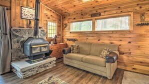 A cozy cabin interior with a stone fireplace, wooden walls, a sofa, and a window with blinds.