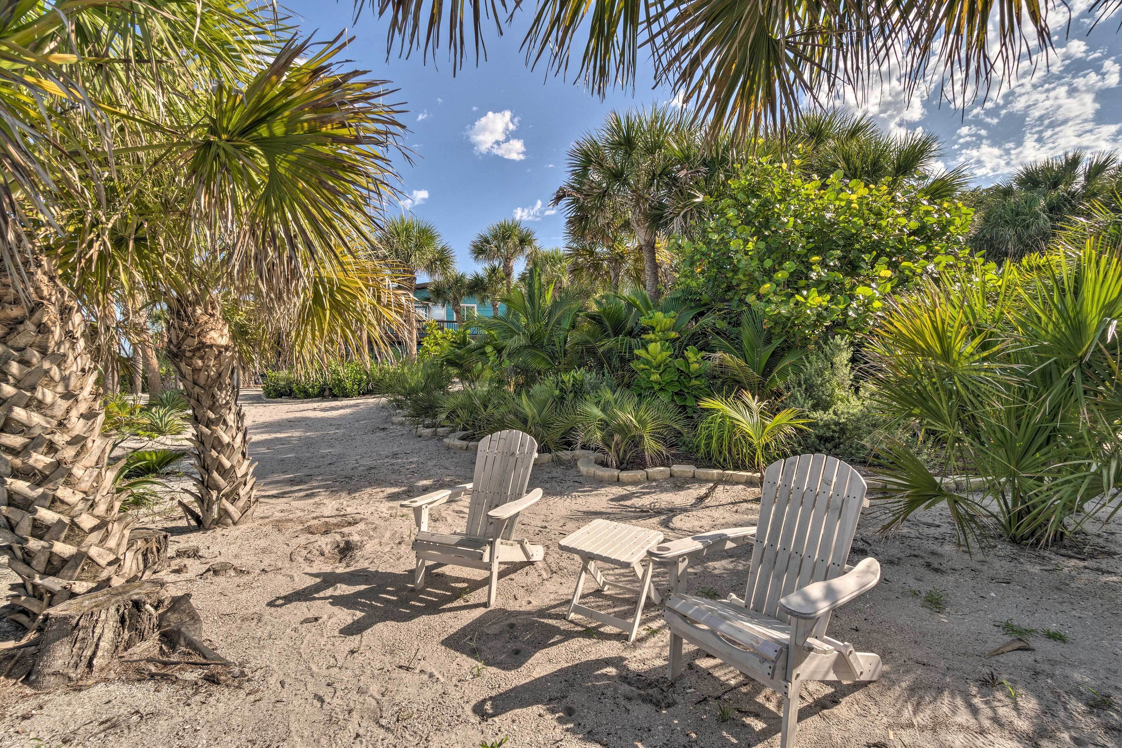 A sandy outdoor area with Adirondack chairs and a small table, surrounded by various tropical plants and palm trees.