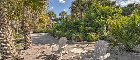 A sandy outdoor area with Adirondack chairs and a small table, surrounded by various tropical plants and palm trees.